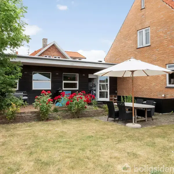 Hus med rødt tag står i en have. Terrasse med havemøbler og parasol, omkranset af blomsterbede med røde blomster. Grønne græsplæne og træer skaber en hyggelig atmosfære.