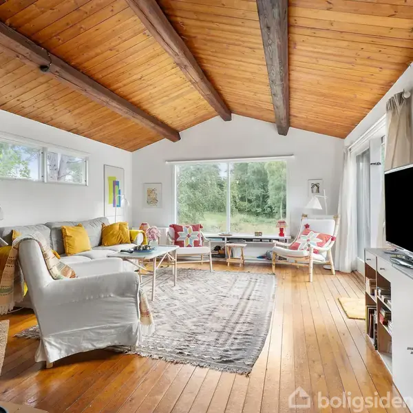 Stue med sofa og lænestole i afslappet arrangement, tæppe på trægulv, og stor vindueskarm med udsigt til frodig have, træbjælker i loftet. Tv står modsat sofaen.