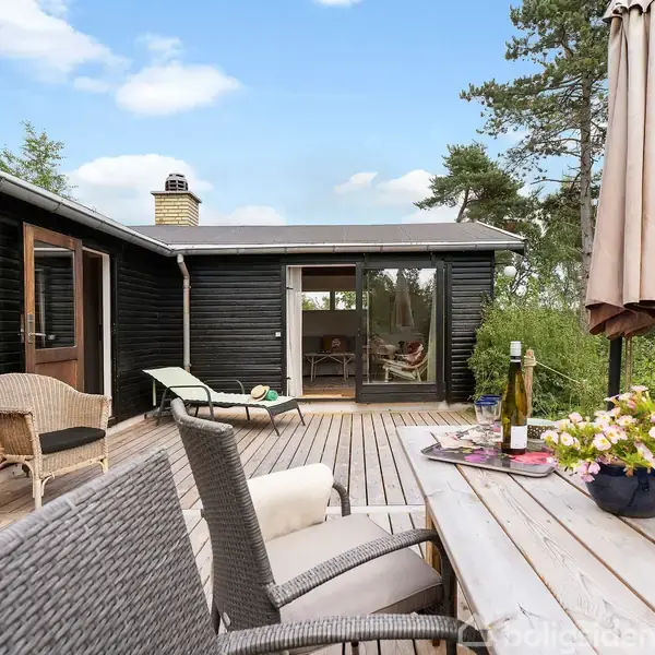 Terrasse med bord og stole, hvorpå der står blomster og en vinflaske, ved et sort træhus. Parasol står slået sammen i tæt omgivelser med skov og himmel i baggrunden.