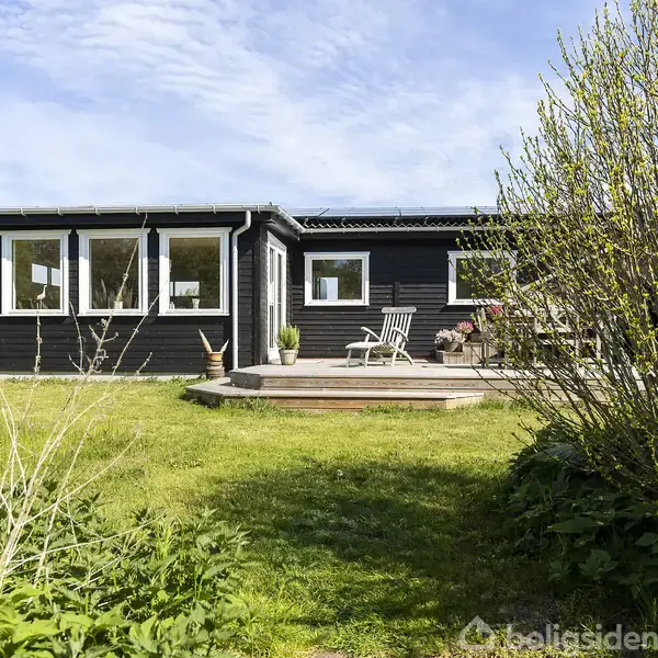 Sort træhus står stille i sollys, omgivet af grøn græsplæne og buske. Foran ses en terrasse med to stole og et bord, hvor planter er placeret.