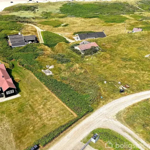Græsareal med flere huse samt en grusvej, der snor sig gennem landskabet. Husene er omgivet af grønne marker og buske nær en kystlinje. Sandstranden kan ses i baggrunden.