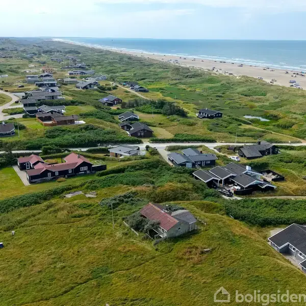 Flere sommerhuse står spredt langs en kystlinje. Husene er omgivet af grønne marker og sandklitter. I baggrunden er der en bred strand ved havet.