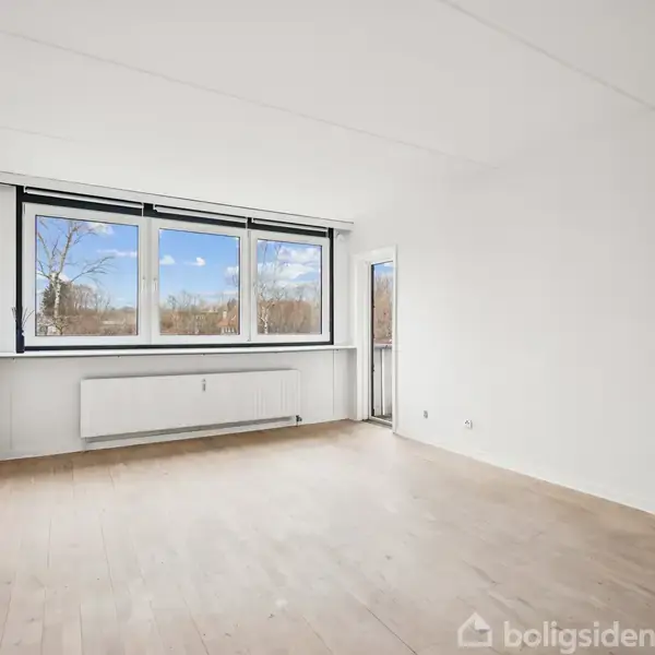 En tom, lys stue med trægulv har et stort vindue, der vender ud mod et træfyldt område. Væggene er hvide, og der er en radiator under vinduet.