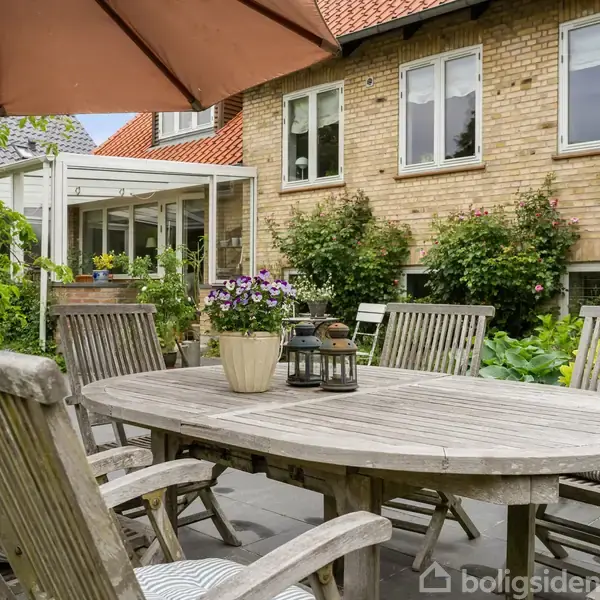 Et træbord med seks stole står på en terrasse under en parasol. En potte med blomster og en lanterne pryder bordet. I baggrunden ses et murstenshus med vinduer og en have med grønne buske.