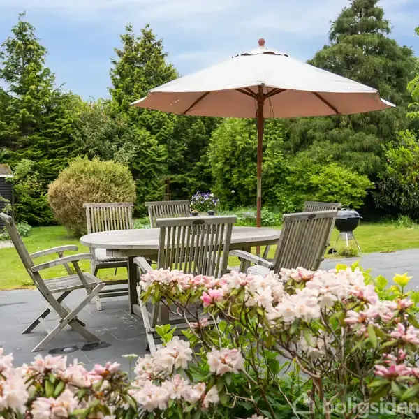 Et havebord med seks stole står på en terrasse under en hvid parasol. Foran er blomstrende buske, og baggrunden består af en frodig have med træer og en grill.