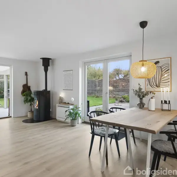 Spisebord med stole står i lyst rum ved siden af et brændekomfur. Store vinduer giver udsigt til en grøn have. Til venstre åbner et rum mod udestue med planter.