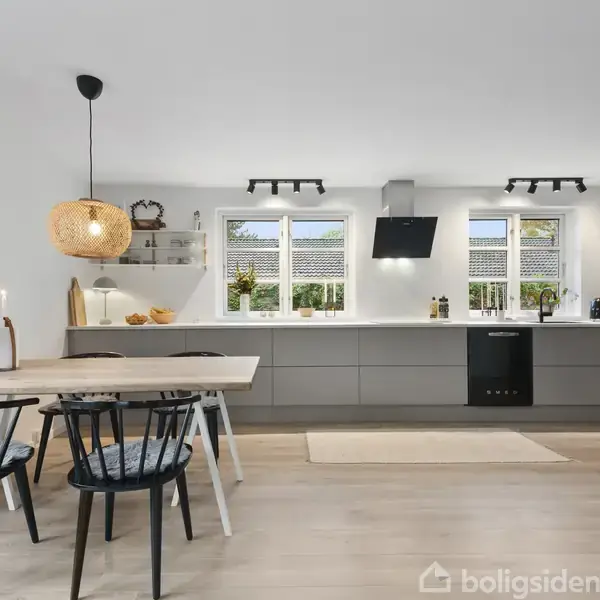 Et moderne køkken-alrum med spisebord og fire stole står i forgrunden. Køkkenet i baggrunden har grå skabe, lys træbordplade og to vinduer, der slipper dagslys ind.