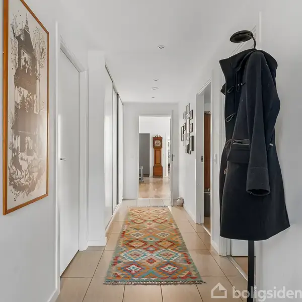 Frakke hænger på en stumtjener i en lysegrå gang med beige fliser. En farverig løber strækker sig langs gulvet, og en stor, gammeldags ur står for enden af gangen.