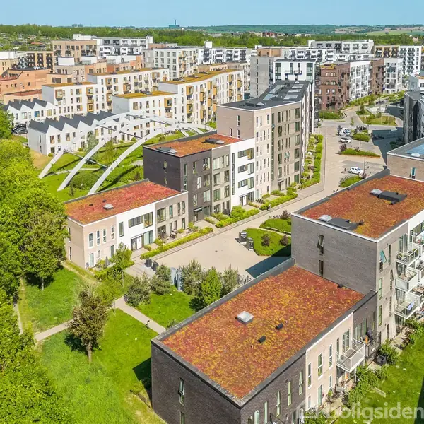 Flere moderne boligbygninger med røde tage står i række i et grønt boligområde, omgivet af træer og græsplæner. Vy af et større byområde med varierende byggehøjder.
