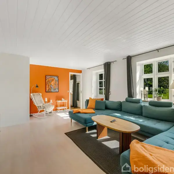 En stue med en blå sofa og orange puder. Ved siden af står et ovalt sofabord. Vægge er lyse, med en enkelt orange væg. Store vinduer giver udsigt til haven.