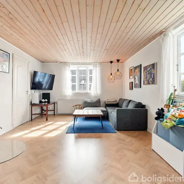 Brændeovn i hjørnet står uden aktivitet i en lys stue med trægulv og loft. Omkring er en sofa, sofabord, væghængt TV og børnelegetøj nær vinduet.