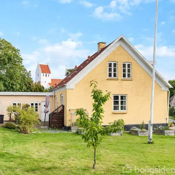 Gul murstenshus med rødt tag står i en grøn have. En flagstang rejser sig ved siden af huset. I baggrunden ses træer og en kirke med et hvidt tårn.