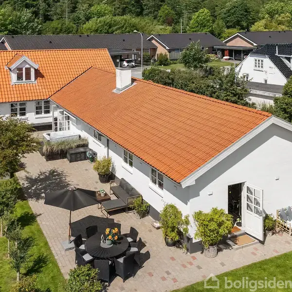 Hvid villa med orange tegltag; rummelig terrasse med møbler og parasol. Omgivet af velplejet have i et roligt boligområde med flere lignende huse i baggrunden.