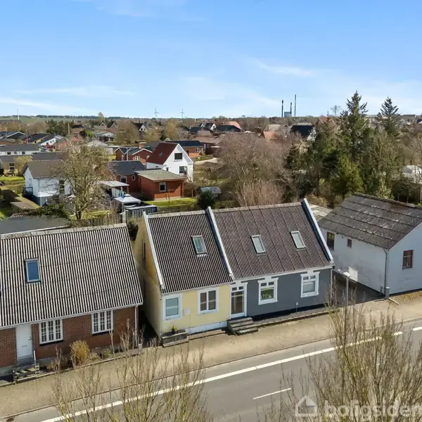 Rækkehuse ligger side om side langs en vej. Husene har forskellige farver og tagformer. I baggrunden ses flere huse og træer under en blå himmel.
