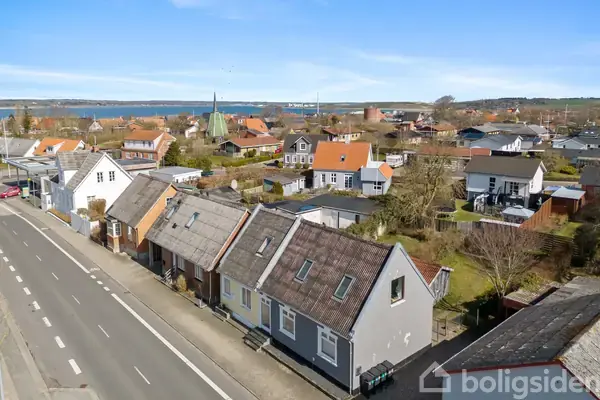 En række huse med skrå tage ligger langs en vej. Landsbyens huse omgiver dem, med en kirke i baggrunden og en fjord i horisonten under en klar himmel.