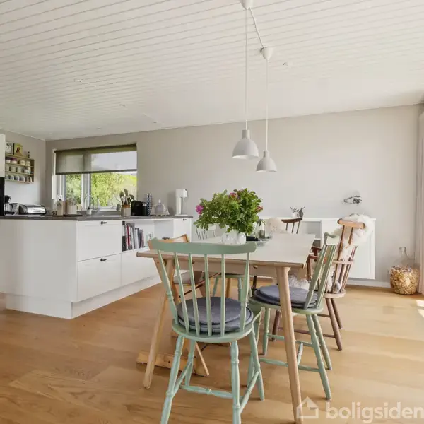 Spisebord med stole står i et lyst, moderne køkkenalrum. Bordet har en vase med blomster. Store vinduer til højre giver naturligt lys. Trægulv og hvidmalede vægge skaber en varm atmosfære.
