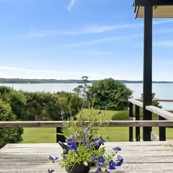 Et bord på en terrasse med blå blomster i en vase. Udsigt til en frodig have og en rolig sø i baggrunden under en klar blå himmel.