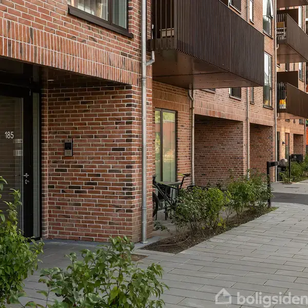 Rød murstensbygning med dør nummer 185 fører til en flisebelagt gangsti. Cykler står op ad væggen, omgivet af grønt græsplæne og buske.