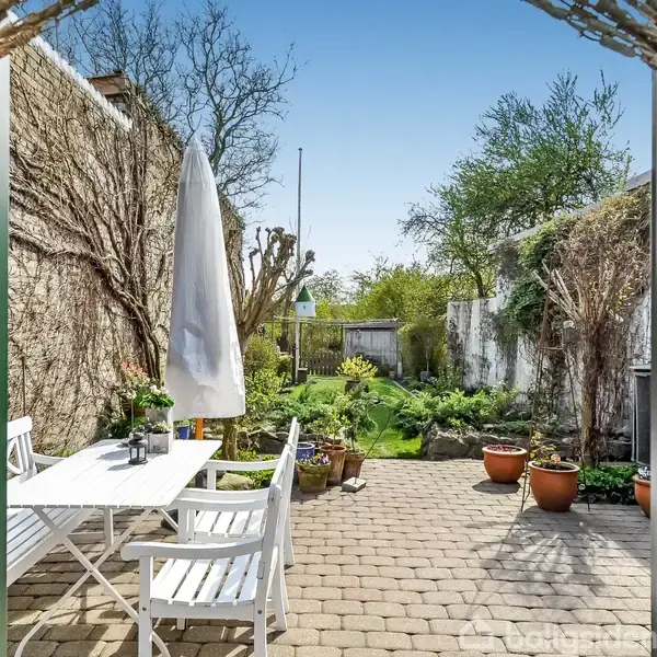Udendørs terrasse med hvidt havemøbelssæt under en stor parasol. Omkring står potteplanter, og der er udsigt til en frodig have med træer og buske.