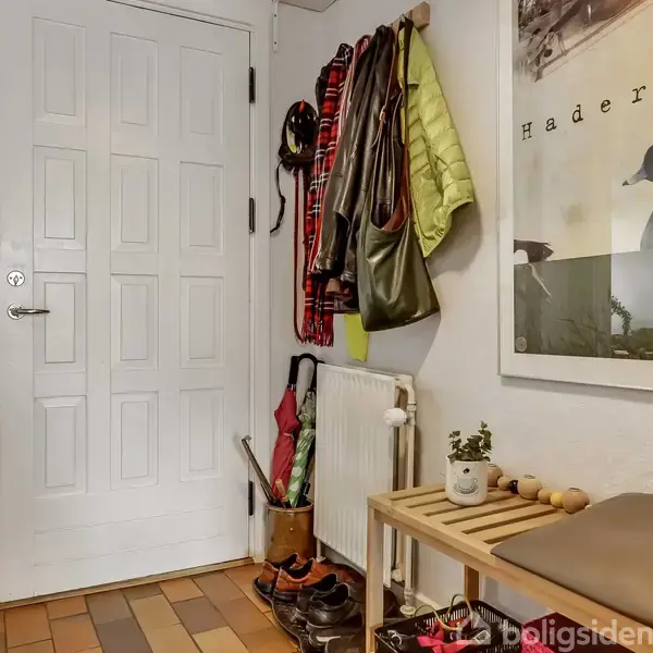 En entre har sko og tasker under en bænk. Jakker hænger på kroge ved siden af en radiator. En plakat med teksten "Haderslev" hænger på væggen.