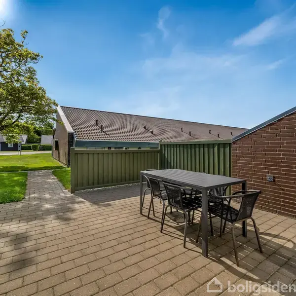 Et udendørs terrassebord med fire stole står på en brolagt terrasse. I baggrunden ses en stor, grøn græsplæne, nogle træer og flere bygninger under en klar blå himmel.