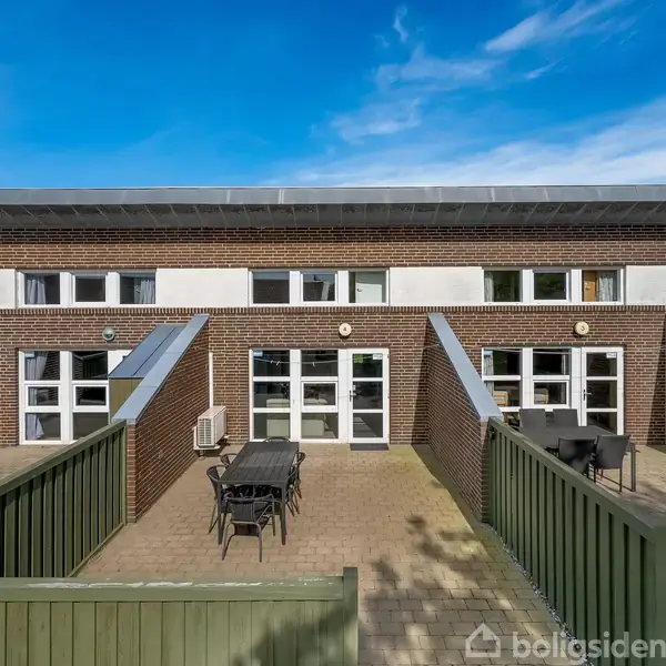 Bygning med mursten udgør baggrunden, hvor terrasser med borde og stole er placeret foran glasdøre. Grønne hegn adskiller rummene, og himlen er klar og blå.