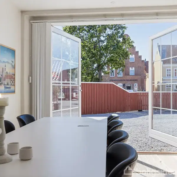 En spisestue med et langt hvidt bord og sorte stole. Døre står åbne mod en gårdhave med grus og en rød mur, omgivet af huse og træer i baggrunden.