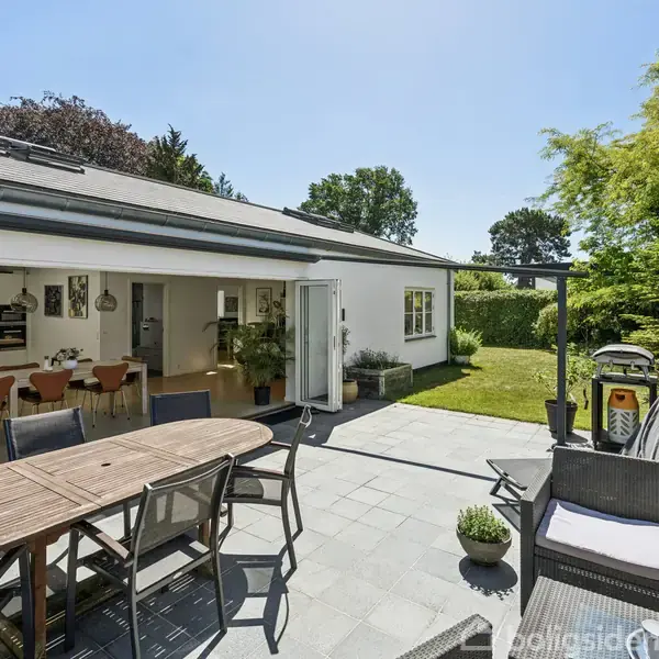 Terrasse med spisebord og stole foran et moderne hus med åben dør til spisestue. Have med grønne buske og græs omkranser området; grill og havemøbler i hjørnet.