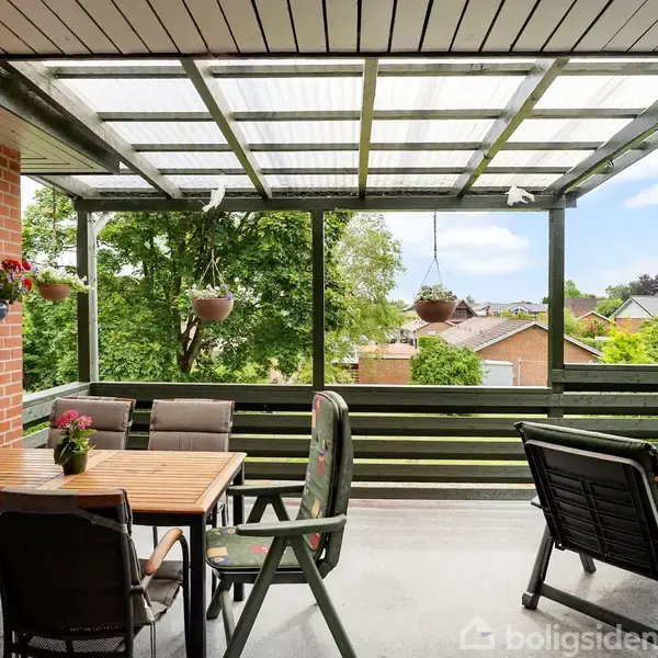 Terrasse med havemøbler er dækket af et gennemsigtigt loft. Den er omgivet af grønne planter og blomster i potter og hængende kurve. Udsigt til huse og træer i nabolaget.