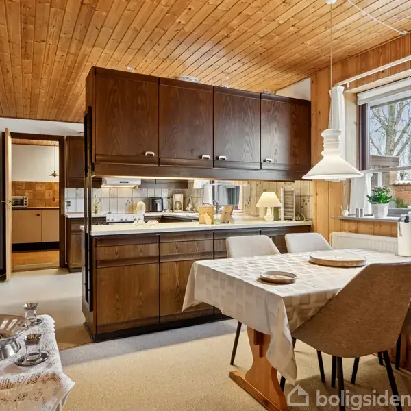 Spisebord står dækket i en hyggelig spisestue med træloft og vægge. Et mørkt skab adskiller spiseområdet fra køkkenet. Et vindue giver udsigt til haven.