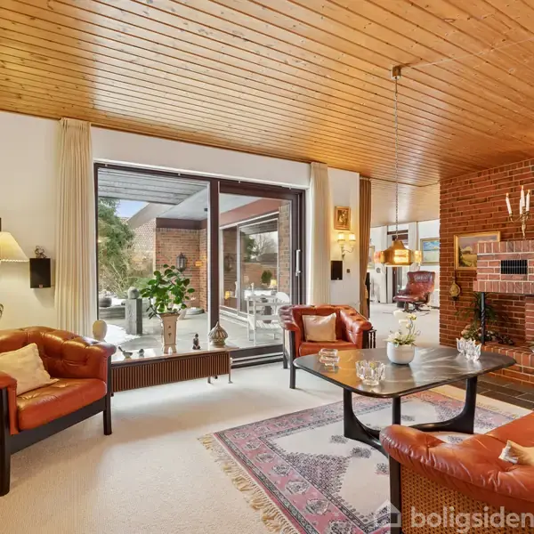 En stue med lædersofaer og et sofabord, foran en murstenspejs med lys. En glasdør fører ud til en terrasse med planter. Bogreoler fylder hjørnet.