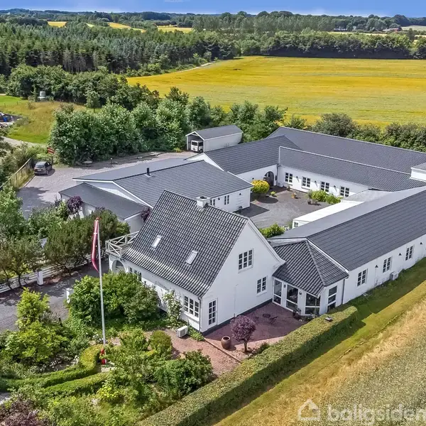 En stor hvid bygning med mørkt tag ligger i et landligt område med marker og skov i baggrunden. En flagstang står foran huset omgivet af grøn have.