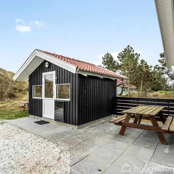 Sort træhus med hvide vinduesrammer og rød tegltag står på flisebelagt terrasse. Træbord/bænkesæt ved siden af huset. Omgivet af klitlandskab med græs og træer.