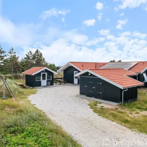 Et sortmalet træhus med rødtag står omgivet af græsbevoksede klitter. Der er en grusvej foran, og en gynge til venstre indikerer en hyggelig, naturskøn beliggenhed.