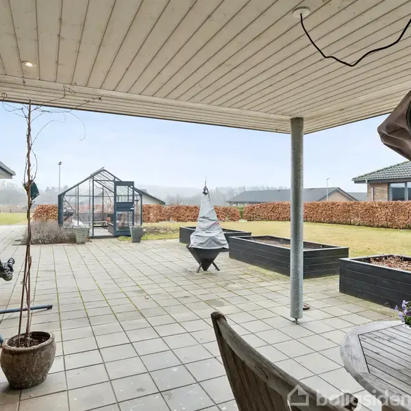 Terrasse med spisebord og stole under overdækning. I baggrunden ses en have med drivhus og forhøjet blomsterbede. Omgivelserne inkluderer flere huse og en hæk.