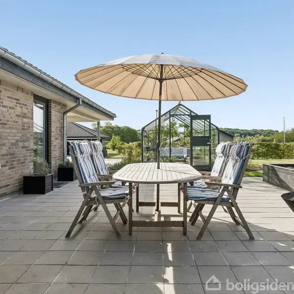 Terrasse med bord og fire stole under en parasol. En grill står nær huset. Bålsted med ild i hjørnet. I baggrunden ses græsplæne og drivhus.