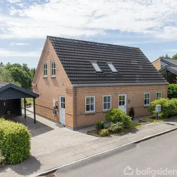 Et rødstenshus med mørkt tegltag og flere vinduer står ved siden af en carport. Forhaven har velplejede buske og blomster. Solrig himmel og skov baggrunden.