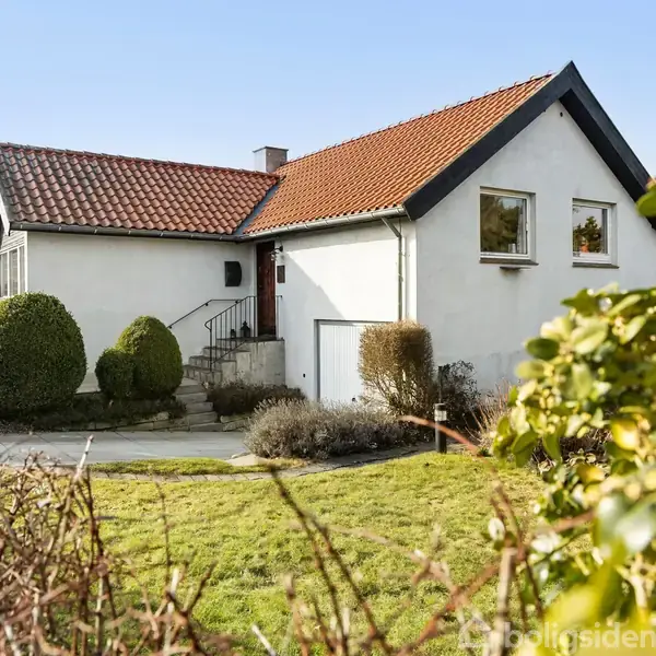 Hvidt hus med rødt tegltag, omgivet af grøn have med buske. Huset har en indgangstrappe og flere vinduer. Klare blå himmel i baggrunden.