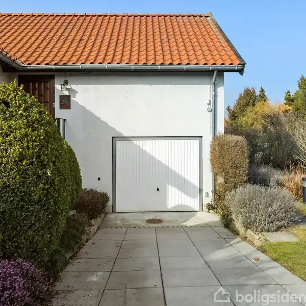 Garage med hvid port beliggende i en have med flisebelagt indkørsel og grøn vegetation. Omkringliggende buske og træer omkranser området, der har et klassisk, boligområdepræg.