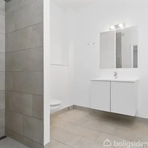 Brusebad i moderne badeværelse med grå flisevægge; vask med hvidt skab og spejl oplyst af væglampe i lysrum. Toilettet er delvist synligt bag en væg.