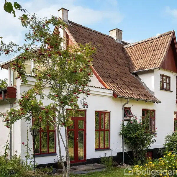 Hvidt hus med rødt tegltag og røde vinduer står i en frodig have med grønne buske og et lille træ. Klar himmel i baggrunden.