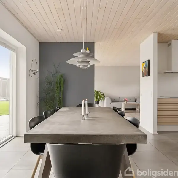 Et langt spisebord med sorte stole er centralt placeret i en lys stue. Store vinduer åbner ud til en grøn have. Modernistisk indretning med minimalistiske træk og grå vægge.