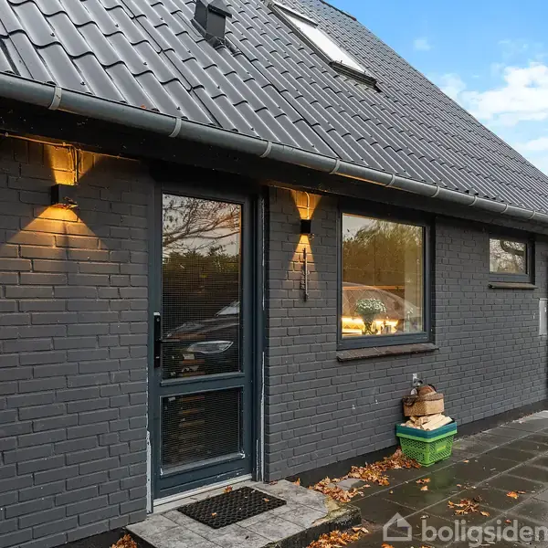 Sort murstenshus med skråt tag og flere vinduer. Udendørs lamper lyser væggen. Fliser dækker jorden foran. Træer ses i baggrunden. Ingen tekst til stede.