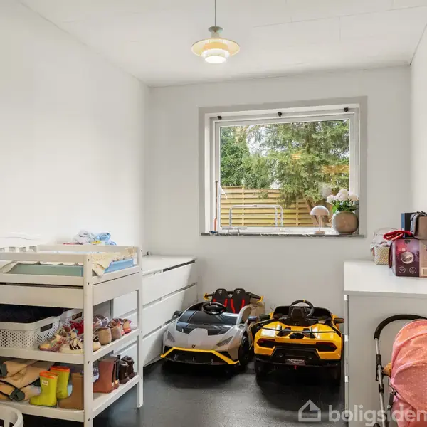 Et lille rum med legetøjsbiler og en barnevogn på gulvet. En kommode står til venstre med opbevaring. Vinduet viser en terrasse og grønne omgivelser udenfor.