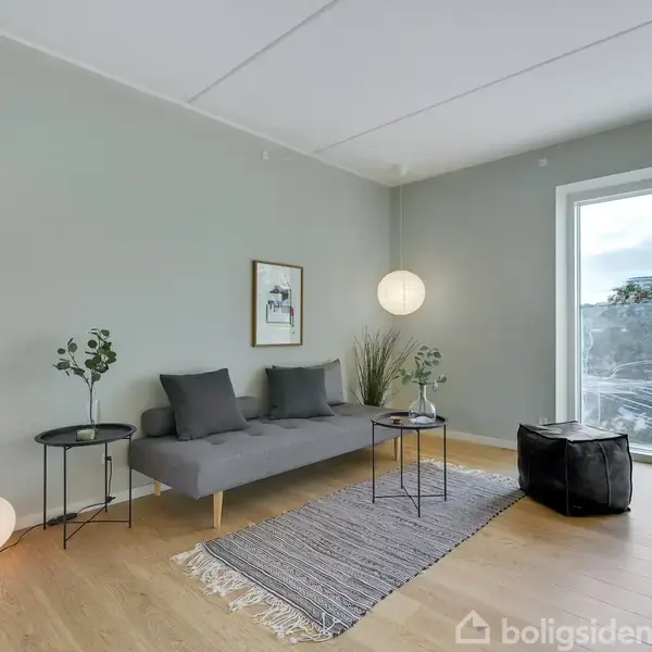 Grå sofa står på et trægulv med en lille tæppe foran. En rund lampe hænger ved siden af. En stor rude viser udsigt over bygninger.