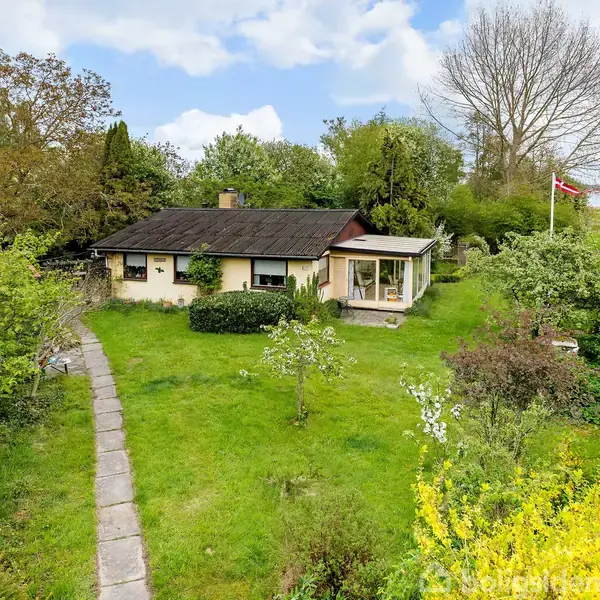 Hus omgivet af grøn have. En flisebelagt sti fører til verandaen. Træer og buske omkranser huset, som ligger ved en mark med en dansk flagstang i baggrunden.