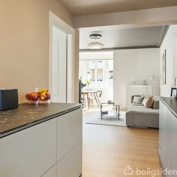 Køkken med mørke bordplader og apparater; frugtskål til venstre. Åbning fører til stue med grå sofa, sofabord og gulvlampe. Vinduer i baggrunden oplyser rummet.