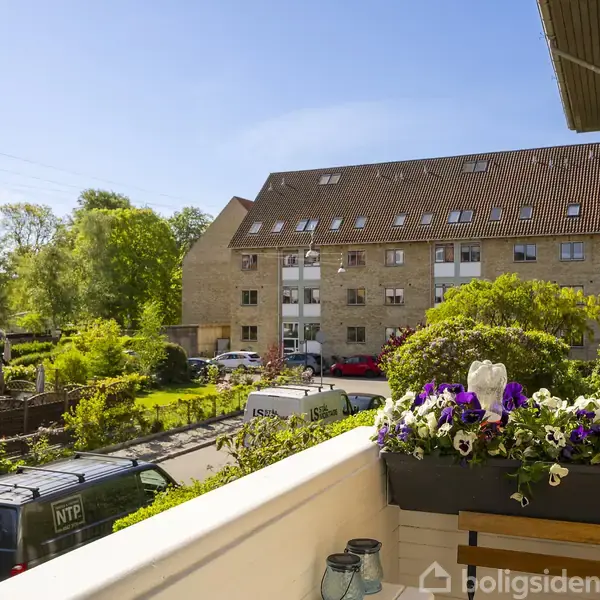 En altan med blomsterkasse fyldt med farverige blomster. Udsigten viser en grøn gårdhave og en række fleretagers bygninger i solskin. To biler er parkeret langs vejen.