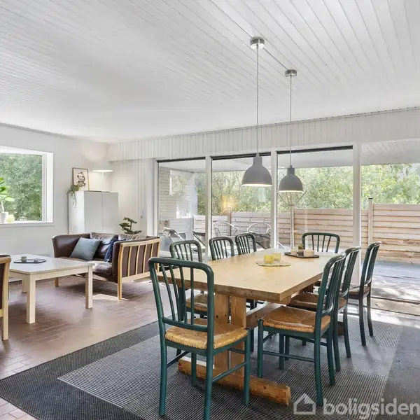 Spisestue med træbord omgivet af otte stole, oplyst af to hængelamper. I baggrunden et opholdsområde med sofa, sofabord, og vindue med udsigt til terrasse og grønne omgivelser.