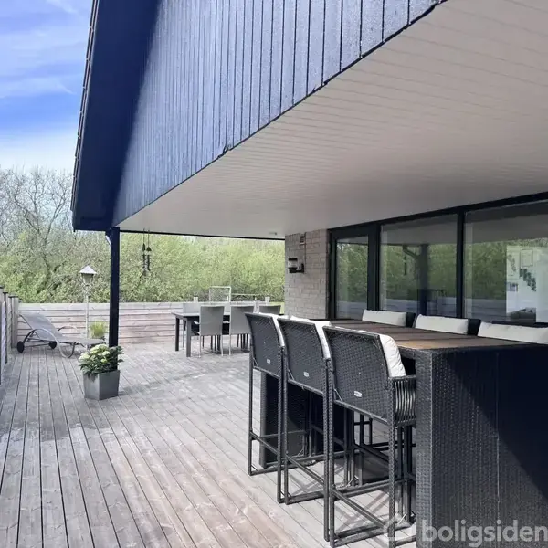 Terrassen har en lang bordplads med høje stole og glaspartier, omgivet af træplanker. I baggrunden ses en spiseplads med bord og stole, skov i horisonten.